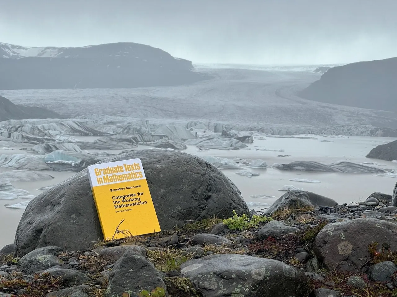 The yellow book on a rounded boulder above a misty meltwater lagoon, a broad grey glacier tongue descending between dark ridges at Hoffellsjökull